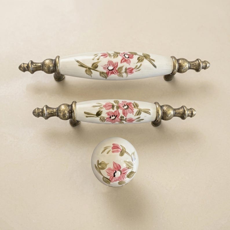 May include: Two elongated drawer pulls and one round cabinet knob made of white ceramic with hand-painted pink flowers and green leaves. The hardware features antique brass-toned metal ends with a decorative, textured, and weathered finish.