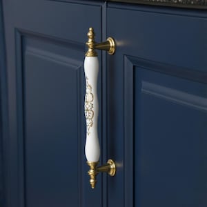 Pode incluir: Uma puxador de armário branco e dourado com um design decorativo. A puxador é montada em uma porta de armário azul escuro. A ferragem tem um acabamento dourado e uma puxador de porcelana branca com detalhes dourados.