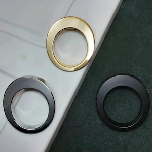 May include: Three circular cabinet pulls in gold, silver, and black. The pulls are arranged on a white and dark green surface. The gold pull is at the top, the silver pull is in the middle, and the black pull is at the bottom.
