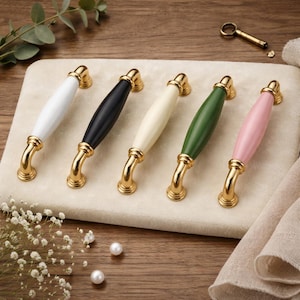 Op de afbeelding: Vijf kastgrepen met goudkleurige hardware en kleurrijke keramische handgrepen. De handgrepen zijn wit, zwart, crème, groen en roze. De grepen zijn gerangschikt op een beige oppervlak, met een kleine sleutel en decoratieve elementen op de achtergrond.