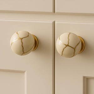 May include: Two cream-coloured cabinet knobs with a gold crackle pattern. The knobs are round and mounted on gold-toned backplates. The cabinet doors are a matching cream colour.