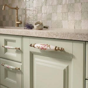 May include: A set of cabinet pulls featuring a white ceramic centre with a hand-painted pink floral design and green leaves, accented by antique brass metal ends. These horizontal handles are mounted on light sage green kitchen cabinet doors.