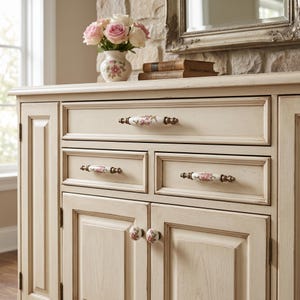 May include: A cream-coloured wooden sideboard featuring a distressed finish, three drawers, and two cabinet doors. The hardware consists of white ceramic handles and knobs decorated with pink floral patterns and antique brass-toned metal accents.
