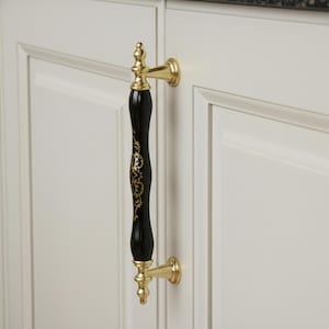 Könnte beinhalten: Ein Schrankgriff mit schwarzem Mittelteil und goldenen Akzenten. Der Griff hat ein dekoratives goldenes Design auf dem schwarzen Mittelteil und ist an einer weißen Schranktür befestigt. Die Hardware ist goldfarben.