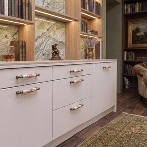 May include: A white cabinet featuring five horizontal drawers and doors with decorative ceramic handles. Each handle is white with a floral pattern of pink roses and green leaves, accented by antique brass-toned metal ends and mounting hardware.