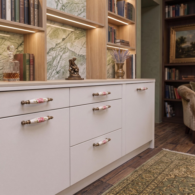 May include: A white cabinet featuring five horizontal drawers and doors with decorative ceramic handles. Each handle is white with a floral pattern of pink roses and green leaves, accented by antique brass-toned metal ends and mounting hardware.