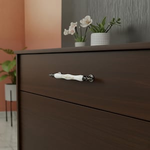 May include: A dark brown wooden chest of drawers featuring a white and gold decorative handle. The handle is porcelain and curved. The chest has two drawers and is in a room with plants and flowers.
