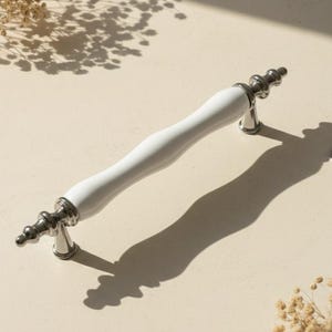 May include: A white ceramic cabinet pull with silver-colored metal accents. The handle has a curved, ergonomic design. The hardware is designed for use on drawers and cabinets.