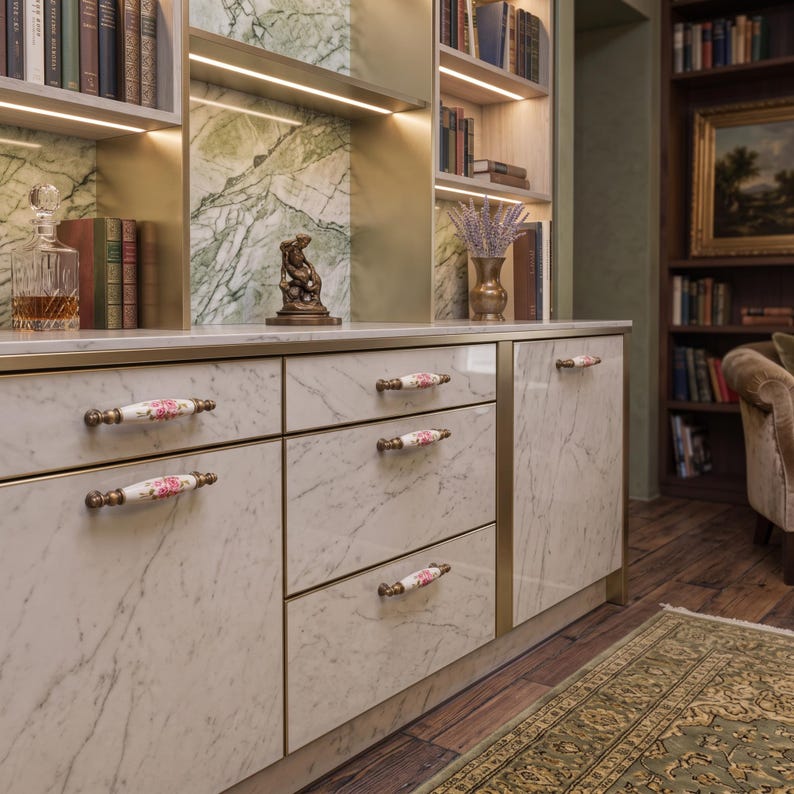 May include: A white marble cabinet featuring five drawers and one door, each equipped with a horizontal ceramic handle decorated with a pink floral pattern and antique brass-toned metal ends, set against a green marble and gold-toned shelving unit.