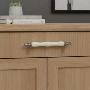 May include: A cream-coloured cabinet handle with silver-coloured detailing, mounted on a light wood cabinet. The handle is a curved shape. The cabinet is made of light-coloured wood with a visible grain.
