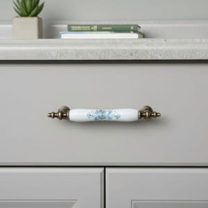 May include: A white and blue floral ceramic cabinet pull with antique brass hardware. The drawer pull is mounted on a light gray cabinet. The cabinet hardware is a decorative accent for kitchen or bathroom furniture.