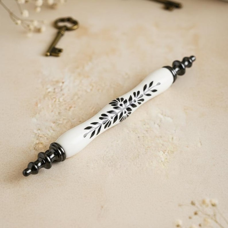 May include: A white cabinet handle with a black floral design. The handle has black accents on each end and a white center with a black floral pattern. This decorative handle is a furniture accessory.