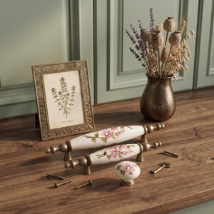 May include: Two white ceramic drawer pulls and one matching round knob, each featuring a hand-painted pink floral design with green leaves and antique brass metal hardware. The set includes several small metal screws and a tiny decorative key.
