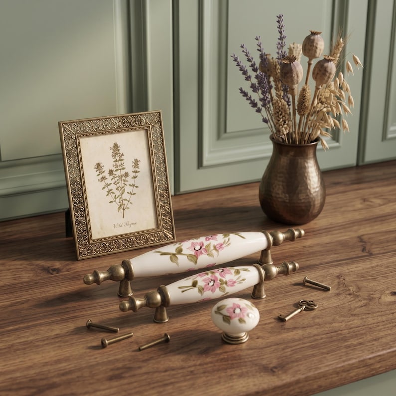 May include: Two white ceramic drawer pulls and one matching round knob, each featuring a hand-painted pink floral design with green leaves and antique brass metal hardware. The set includes several small metal screws and a tiny decorative key.