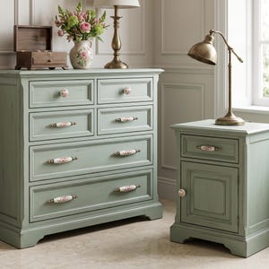 May include: A sage green wooden chest of drawers with four drawers and a matching bedside table with one drawer and one cupboard door. Both pieces feature white ceramic knobs and handles decorated with pink floral patterns and brass-toned metal hardware.