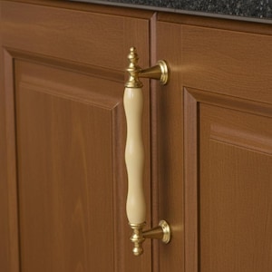 May include: A cream and gold cabinet handle attached to a brown wooden door. The handle has a curved, cream-coloured central section and gold-coloured metal fittings. The design is traditional.