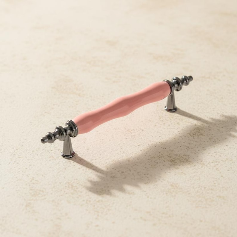 May include: A cabinet pull with a light pink, ridged centre and dark grey metal ends. The decorative hardware has a classic design and is suitable for drawers or cabinets. The pull is approximately 15 cm long.