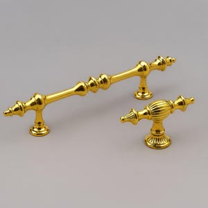Könnte beinhalten: Ein polierter goldfarbener Schrankgriff aus Metall und ein passender Schrankknopf. Der Griff verfügt über eine lange, schlanke Stange mit dekorativen spindelartigen Details und zwei Befestigungssockeln. Der Knopf hat eine gerippte ovale Form.