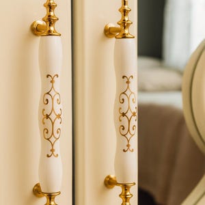 May include: Two white cabinet handles with gold detailing. The handles feature a curved design with gold filigree patterns and ornate gold bases and tops. They are mounted on a cream-coloured cabinet door.