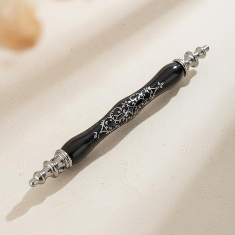 May include: A black and silver decorative pen with a detailed floral design. The pen has a glossy black finish with silver accents at each end and along the barrel. The pen is approximately 18 cm long.
