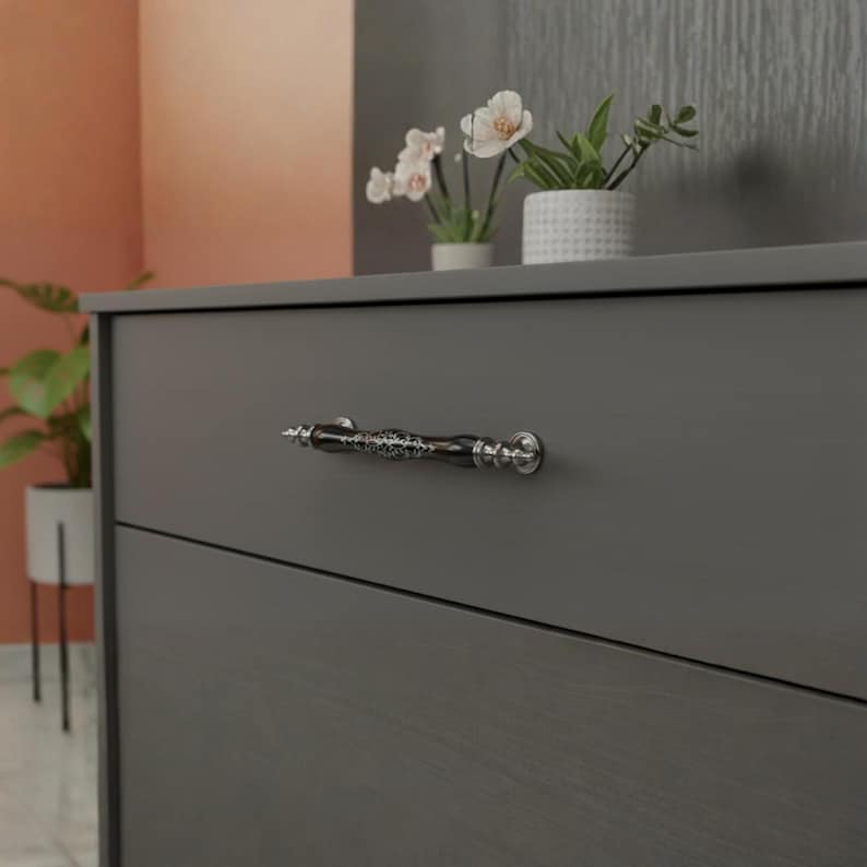 May include: A dark grey chest of drawers featuring a decorative handle. The handle is black with silver detailing and an intricate pattern. The chest has two drawers and is set against neutral-toned walls and decorative plants.