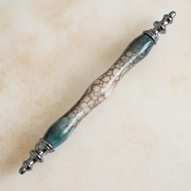 May include: A decorative pen with a marbled effect in teal, grey, and white. The pen has dark metal ends. The pen's body has a unique pattern. The pen is designed for writing or as a decorative item, adding a touch of elegance.