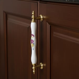 May include: A white porcelain cabinet pull with a floral design in purple, yellow, and blue. The handle is accented with gold-colored metal hardware and is mounted on a dark brown cabinet door. The pull is a decorative element for kitchen or bathroom cabinetry.