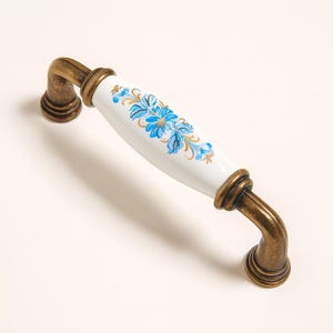 Op de afbeelding: Een kastgreep met een wit keramisch middenstuk met een blauw bloemmotief en gouden accenten. De gebogen handgrepen en de basis zijn gemaakt van antiek messingkleurig metaal. Meubelbeslag.