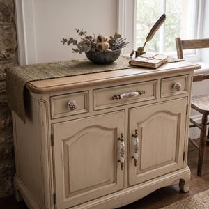 May include: A light beige wooden sideboard featuring a natural wood top, two cabinet doors, and three drawers. The furniture is accented with white ceramic knobs and handles decorated with floral patterns in pink, green, and gold metallic hardware.