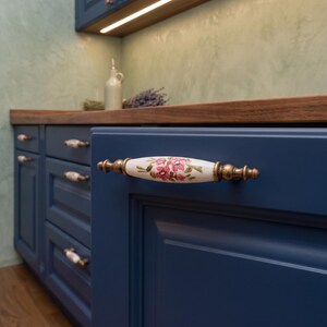 May include: A cabinet handle featuring a white ceramic centre with a pink floral pattern and green leaves, flanked by antique brass-toned metal ends. The handle is mounted on a dark blue cabinet door, showcasing a classic design for furniture hardware.