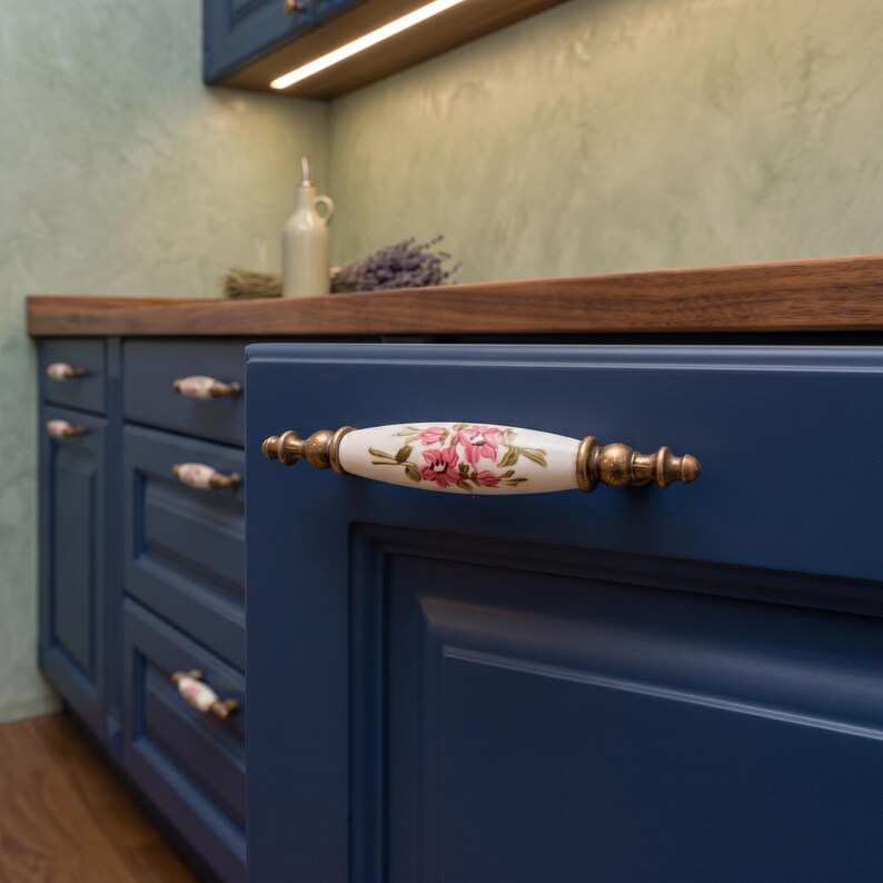 May include: A cabinet handle featuring a white ceramic centre with a pink floral pattern and green leaves, flanked by antique brass-toned metal ends. The handle is mounted on a dark blue cabinet door, showcasing a classic design for furniture hardware.