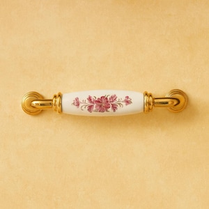 Op de afbeelding: Een decoratieve kastgreep met een wit keramisch middenstuk met een bloemmotief in roze en paarse tinten. De uiteinden en accenten zijn goudkleurig, wat een elegante uitstraling creëert. De greep is ontworpen voor eenvoudige installatie.