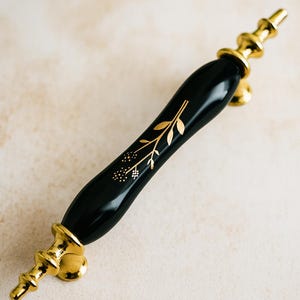 May include: A black cabinet pull with gold detailing. The handle has a glossy black finish with a gold floral pattern. The ends and base are gold, creating a decorative hardware piece for furniture.