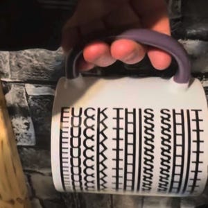 Puede incluir: Una taza de cerámica blanca con un asa morada que presenta el texto negro repetido "FUCK THIS SHIT" impreso en una orientación vertical y apilada en el lateral de la taza, creando un patrón visual distorsionado y repetitivo.