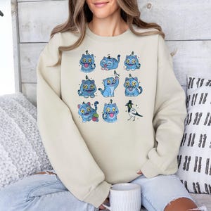 May include: A cream-colored sweatshirt featuring a repeating pattern of blue cartoon cats with yellow eyes and a black bird. The cats have various expressions and poses. The sweatshirt is made of a soft material.
