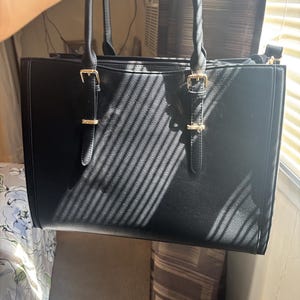 May include: A black tote bag with gold-colored hardware, including buckles and zippers. The bag has two top handles and a shoulder strap. The material appears to be leather or a leather-like material. The bag is rectangular and has a structured design.