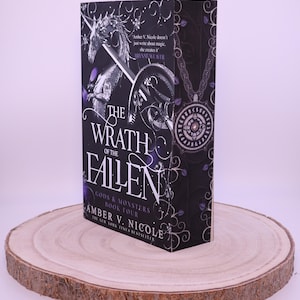 Peut inclure: Un livre &agrave; couverture rigide intitul&eacute; "The Wrath of the Fallen" par Amber V. Nicole. Le livre pr&eacute;sente une couverture noire avec des accents argent&eacute;s et violets, et un dessin d'une licorne et d'une &eacute;p&eacute;e. Le livre est pr&eacute;sent&eacute; sur une tranche de bois ronde.