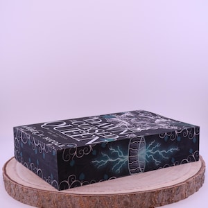 Peut inclure: Une bo&icirc;te rectangulaire avec le titre "The Cursed Queen" en lettres blanches. La bo&icirc;te a un fond sombre avec des accents turquoise et un graphique d'un portail avec des &eacute;clairs. La bo&icirc;te repose sur une tranche de bois.