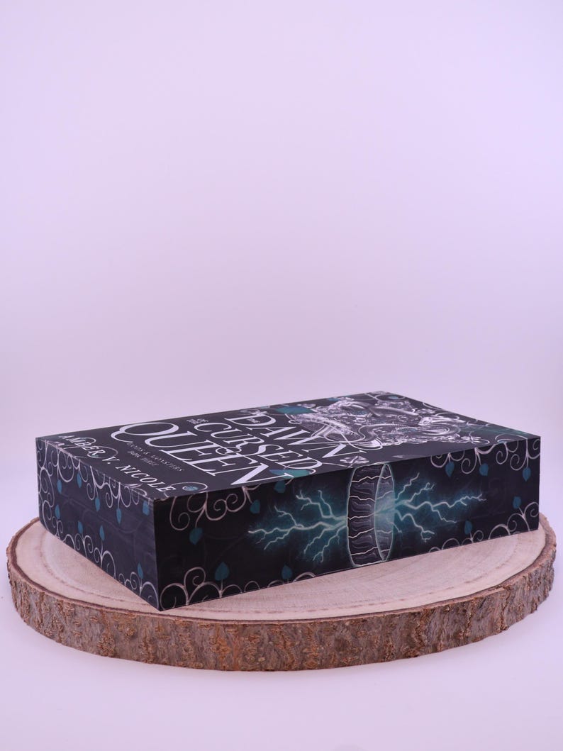 Peut inclure: Une bo&icirc;te rectangulaire avec le titre "The Cursed Queen" en lettres blanches. La bo&icirc;te a un fond sombre avec des accents turquoise et un graphique d'un portail avec des &eacute;clairs. La bo&icirc;te repose sur une tranche de bois.