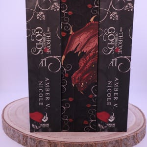 Peut inclure: Trois livres &agrave; couverture rigide intitul&eacute;s "The Throne of Broken Gods II" par Amber V. Nicole. Les dos sont noirs avec du texte blanc et des motifs d&eacute;coratifs. Le livre central pr&eacute;sente une illustration de dragon rouge. Les livres sont pr&eacute;sent&eacute;s sur une tranche de bois.