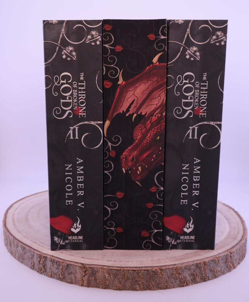 Peut inclure: Trois livres &agrave; couverture rigide intitul&eacute;s "The Throne of Broken Gods II" par Amber V. Nicole. Les dos sont noirs avec du texte blanc et des motifs d&eacute;coratifs. Le livre central pr&eacute;sente une illustration de dragon rouge. Les livres sont pr&eacute;sent&eacute;s sur une tranche de bois.