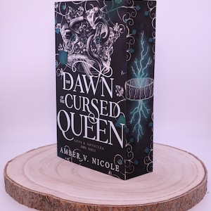Peut inclure: Un livre &agrave; couverture rigide intitul&eacute; "The Dawn of the Cursed Queen" par Amber V. Nicole, avec une couverture noire orn&eacute;e d'une couronne argent&eacute;e et d'un motif de serpent. Le livre est pr&eacute;sent&eacute; sur une tranche de bois ronde.