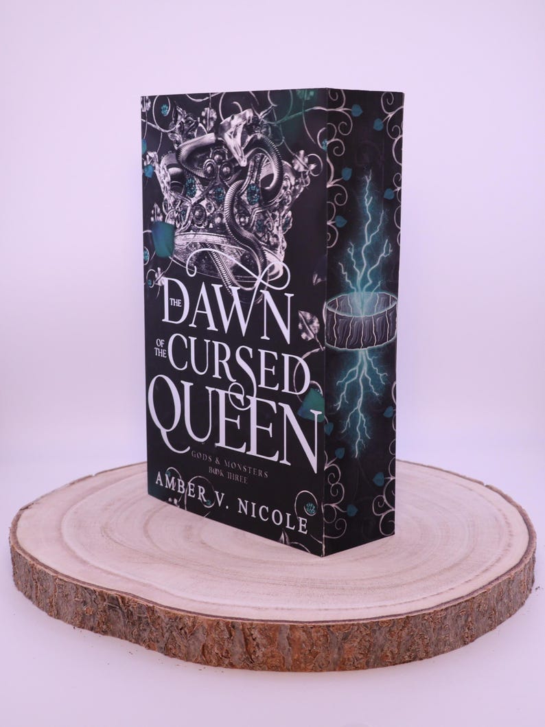 Peut inclure: Un livre &agrave; couverture rigide intitul&eacute; "The Dawn of the Cursed Queen" par Amber V. Nicole, avec une couverture noire orn&eacute;e d'une couronne argent&eacute;e et d'un motif de serpent. Le livre est pr&eacute;sent&eacute; sur une tranche de bois ronde.