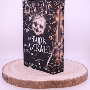 Peut inclure: Un livre &agrave; couverture rigide intitul&eacute; "The Book of Azrael" avec une couverture noire orn&eacute;e d'un cr&acirc;ne, d'une couronne et d'une &eacute;p&eacute;e. Le livre est pr&eacute;sent&eacute; sur une tranche de bois ronde, avec le titre et l'auteur, Amber V. Nicole.