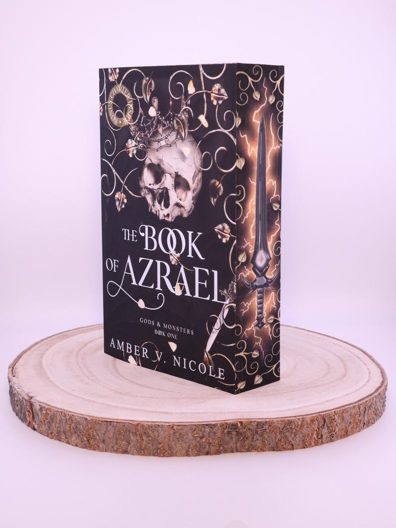 Peut inclure: Un livre &agrave; couverture rigide intitul&eacute; "The Book of Azrael" avec une couverture noire orn&eacute;e d'un cr&acirc;ne, d'une couronne et d'une &eacute;p&eacute;e. Le livre est pr&eacute;sent&eacute; sur une tranche de bois ronde, avec le titre et l'auteur, Amber V. Nicole.
