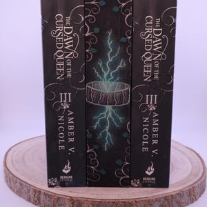 Peut inclure: Trois livres &agrave; couverture rigide intitul&eacute;s "The Dawn of the Cursed Queen" par Amber V. Nicole. Les couvertures des livres sont noires avec des accents blancs et turquoise, avec un motif d'&eacute;clair. Les livres sont pr&eacute;sent&eacute;s sur une tranche de bois.