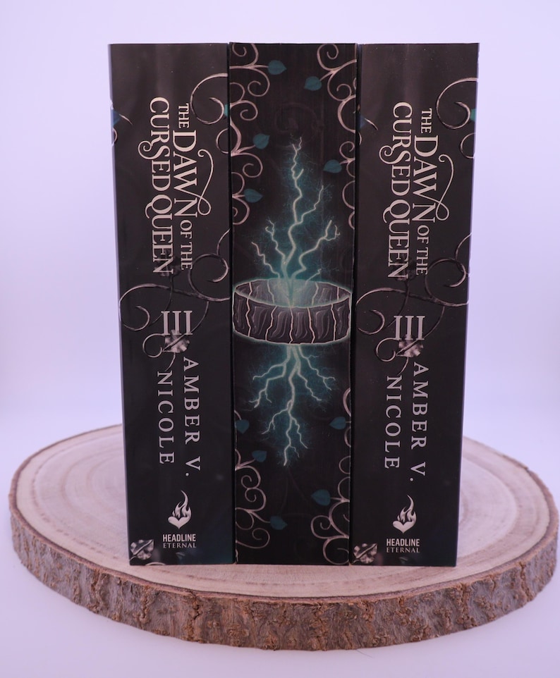 Peut inclure: Trois livres &agrave; couverture rigide intitul&eacute;s "The Dawn of the Cursed Queen" par Amber V. Nicole. Les couvertures des livres sont noires avec des accents blancs et turquoise, avec un motif d'&eacute;clair. Les livres sont pr&eacute;sent&eacute;s sur une tranche de bois.