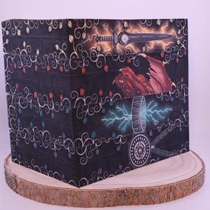 Peut inclure: Une bo&icirc;te d&eacute;corative sur le th&egrave;me de la fantaisie. La bo&icirc;te pr&eacute;sente des illustrations d'une &eacute;p&eacute;e, d'un dragon et d'un marteau avec des effets d'&eacute;clair. La bo&icirc;te est noire avec un motif floral et est pos&eacute;e sur une tranche de bois.