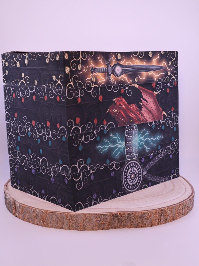 Peut inclure: Une bo&icirc;te d&eacute;corative sur le th&egrave;me de la fantaisie. La bo&icirc;te pr&eacute;sente des illustrations d'une &eacute;p&eacute;e, d'un dragon et d'un marteau avec des effets d'&eacute;clair. La bo&icirc;te est noire avec un motif floral et est pos&eacute;e sur une tranche de bois.