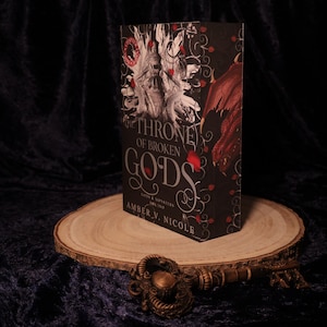 Peut inclure: Un livre intitul&eacute; "Throne of Broken Gods" d'Amber V. Nicole, avec une couverture noire orn&eacute;e de d&eacute;tails rouges et blancs. Le livre est pr&eacute;sent&eacute; sur une tranche de bois, avec une cl&eacute; d&eacute;corative devant. Roman fantastique.